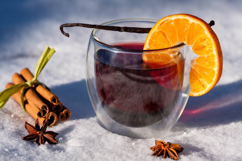 Ein Glas Glühwein mit Orangenscheibe, Vanillestange, Zimt und Nelken steht im Schnee.