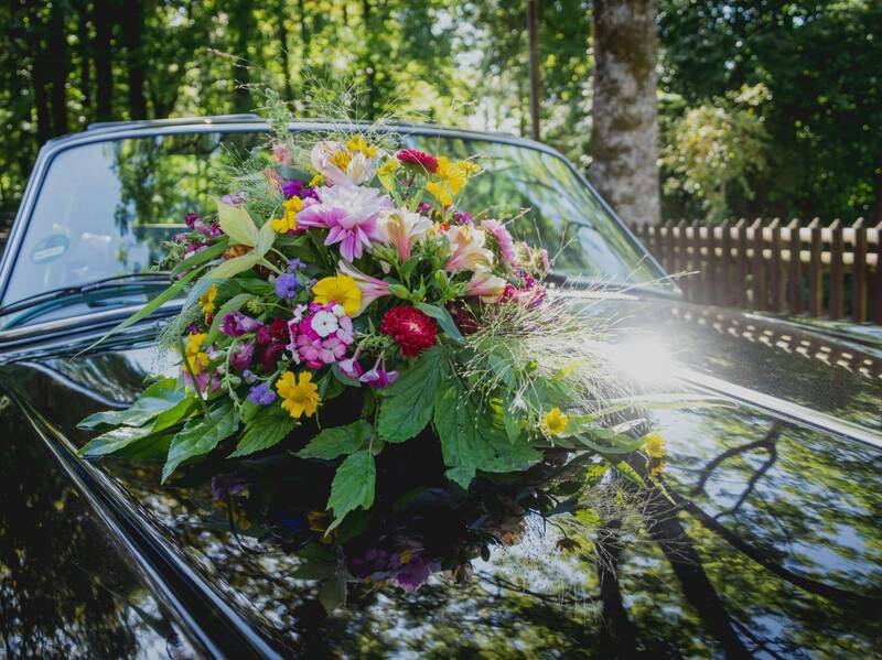 Ein schwarzes Auto, auf dessen Motorhaube ein bunter Hochzeitsblumenstrauß angebracht ist.