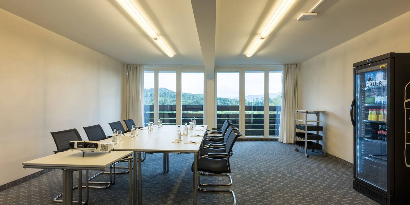 Ein großer Workshopraum mit Tischblock in der Mitte und Stühlen darum herum, an der rechten Wand steht ein Kühlschrank, am Ende des Raums sieht man einen Balkon.