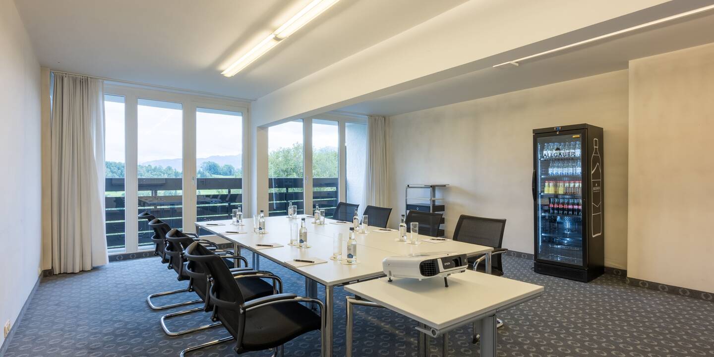 Workshopraum mit eingedeckten Tischblöcken in der Mitte des Raumes, Beamer und Kühlschrank.